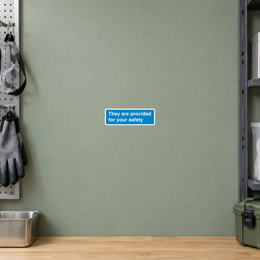 Safety Sign Sticker applied in a smooth sage panel at a general PPE equipment pickup station, safety goggles and gloves on hooks extreme left, central uninterrupted wall