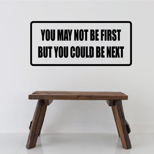 Rectangular wall decal with black border and bold white text: 'YOU MAY NOT BE FIRST'