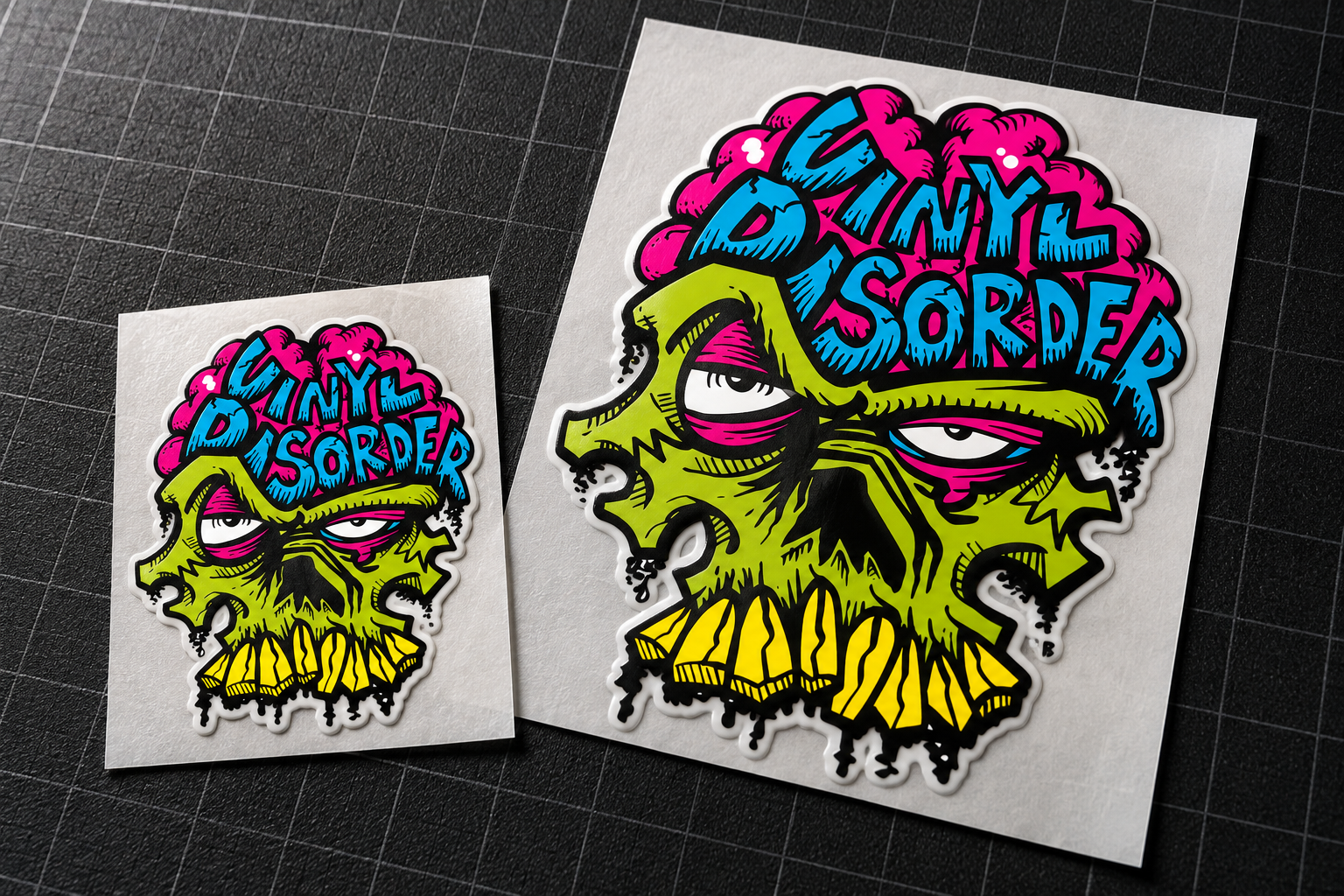 Illustrative photograph-style mockup of two different-size Vinyl Disorder skull stickers on intact liners, on a dark cutting mat.