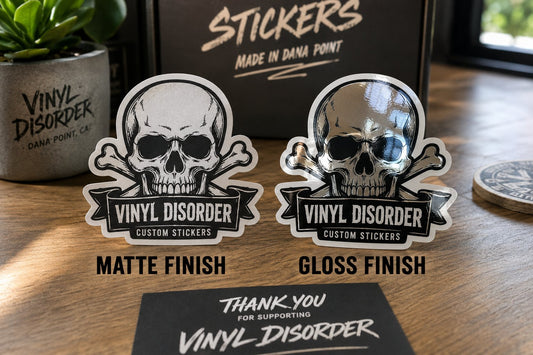 Matte and gloss Vinyl Disorder sticker samples compared side by side on a wooden tabletop under angled light
