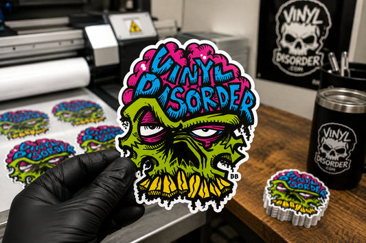 Black-gloved hand holding a finished Vinyl Disorder die cut sticker with a white border in a print shop