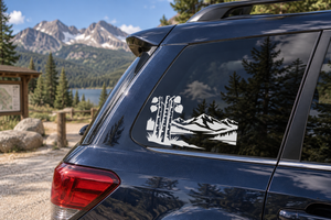 Black-and-white vinyl decal of tall pines, mountains, and lake on car window