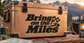 Tan hard cooler box with black handles and “Bring on the Miles” lettering