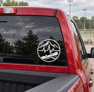 Round vinyl decal with mountain peaks, heartbeat line, and snowboarder boots on red car glass