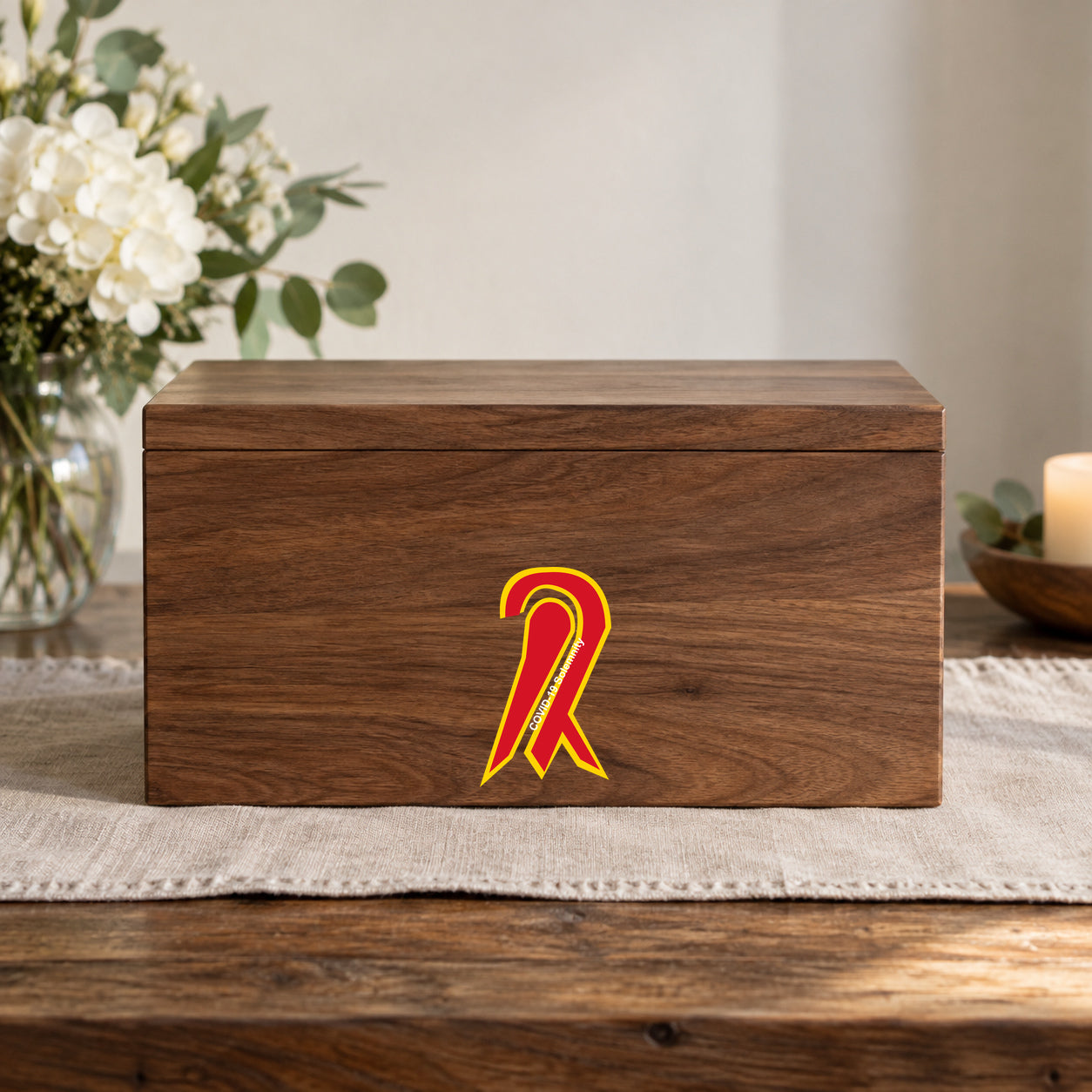 COVID-19 Coronavirus Solemnity Ribbon Vinyl Sticker applied in a smooth sealed walnut memory box front on a remembrance table, flower vase blurred at far left and soft linen beneath, square frontal composition on flat wooden box front