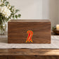 COVID-19 Coronavirus Solemnity Ribbon Vinyl Sticker applied in a smooth sealed walnut memory box front on a remembrance table, flower vase blurred at far left and soft linen beneath, square frontal composition on flat wooden box front