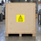 Caution Heavy Load Use Forklift Sticker applied in Large pale plywood shipping crate sitting on a pallet in a machine-tool freight depot, broad flat front board panel without seams or straps in central area, pallet visible beneath and trolley edge far right.