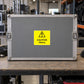 Caution Heavy Sticker applied in Closed broad gray steel freight transport case on a packing bench in a concert equipment warehouse, wide flat central face, latches at extreme edges only, peripheral coiled cable and shelving.