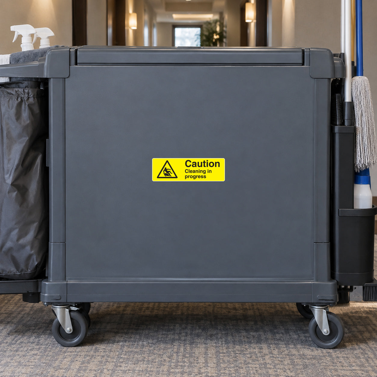 Caution Cleaning In Progress Sticker applied in Front of a wide closed slate-gray commercial janitorial cart in a quiet hotel service corridor, broad flat unbroken plastic front panel central, caster wheels below and cleaning tools only at extreme edges.