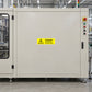 Caution The Machinery Is Automatic Sticker applied in Broad smooth ivory guarded robotic packaging machine enclosure panel in a clean manufacturing hall, central unbroken face, closed inspection window extreme left and controls extreme right.