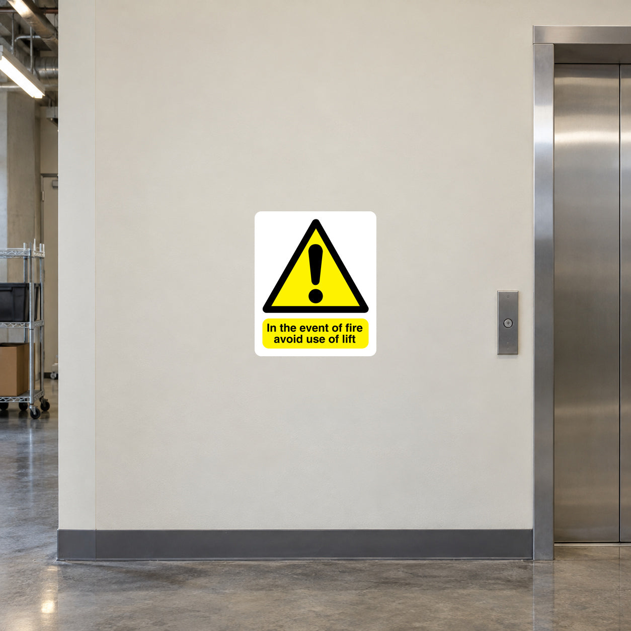 In The Event Of Fire Avoid Use of Lift Sticker applied in Smooth pale cream wall beside a service lift in a commercial building, central panel faces camera unobstructed, lift door at far right and call button outside central region.