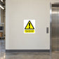 In The Event Of Fire Avoid Use of Lift Sticker applied in Smooth pale cream wall beside a service lift in a commercial building, central panel faces camera unobstructed, lift door at far right and call button outside central region.