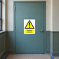 Caution Asbestos Sticker applied in Closed muted teal restricted renovation access door in an older institutional corridor, single opaque flat door central, hardware at far right and protective floor covering below, no exposed debris.
