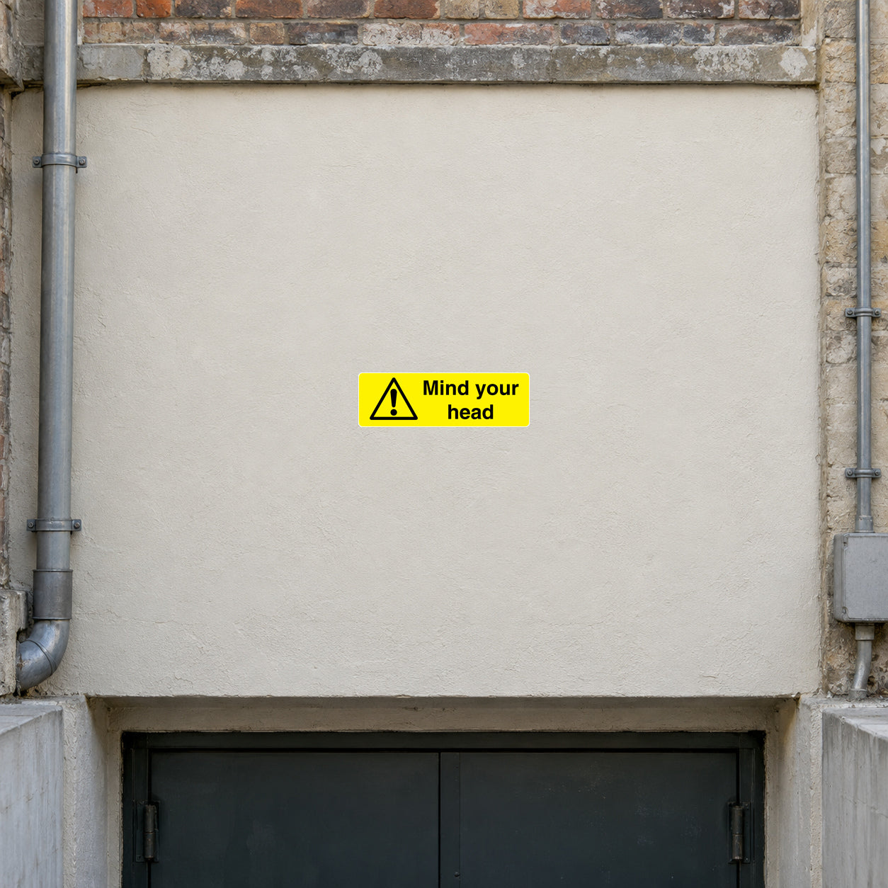 Danger Mind Your Head Sticker applied in Broad smooth flat ivory fascia above a low basement doorway at an old warehouse, central fascia clear and unobstructed across x25%-75% y15%-74%, opening visible only lower edge, pipes far sides.