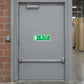 Exit For Emergency Use Only Sticker applied in A solid pale-gray emergency exit door in a paper converting plant, crash bar only at bottom edge, exposed terracotta brick at extreme left and stacked cardboard rolls far right.