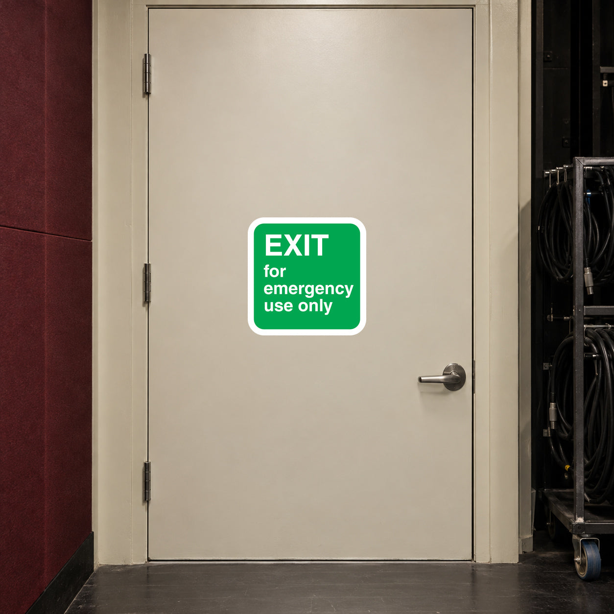 Exit for Emergency Use Only Sticker applied in A smooth cream emergency exit door in a small theatre backstage corridor, maroon acoustic wall at far left, stage cable trolley at far right, handle at bottom right.