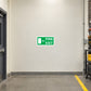 Emergency Fire Exit Over Here Sticker applied in Blank smooth cream wall in a tool hire warehouse corridor, fire exit doorway at extreme LEFT edge, yellow floor stripe and utility cart far right.