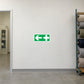 First Aid Shower Sticker applied in Blank smooth off-white corridor wall in a furniture upholstery factory, first aid room doorway at extreme LEFT, an unmarked medical cabinet partly visible inside, textile rack far right.