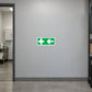 Aid Left Sticker applied in Blank smooth soft gray wall in an electrical workshop staff corridor, first aid room doorway at extreme LEFT, unmarked medical cabinet visible inside, clean concrete floor.