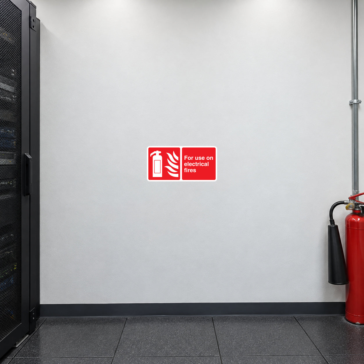 Use On Electrical Fires Sticker applied in a commercial broadcast control equipment room, smooth cool white wall, red CO2 extinguisher with black horn far lower right, closed black equipment rack edge left, raised dark antistatic floor