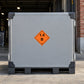 Hazardous Safety Sign Sticker applied in a covered freight departure bay with red brick pier at far right, flat grey rigid cargo case front, dark corner bumpers and pallet bottom