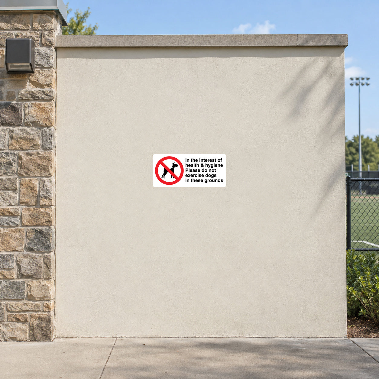 NO Dogs Sign Sticker applied in a smooth ivory rendered recreation-ground perimeter wall, distant sports field glimpsed only at right edge