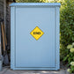 Road Sign Sticker applied in A pale blue rural property maintenance cupboard with path tools extreme left and garden greenery far right, single smooth blank door