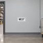 Road Sign Sticker applied in A light grey warehouse wayfinding corridor wall with pallet cart at extreme right and doorway far left, blank smooth center