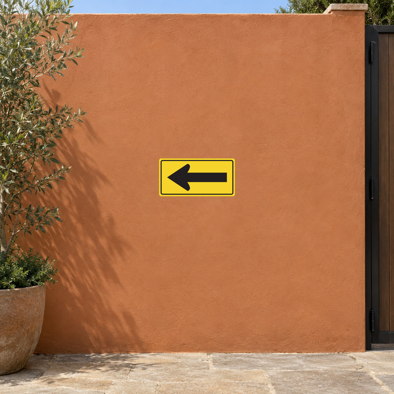 Road Sign Sticker applied in A small industrial yard entrance, uninterrupted terracotta stucco wall, gate edge far right and olive planter bottom left