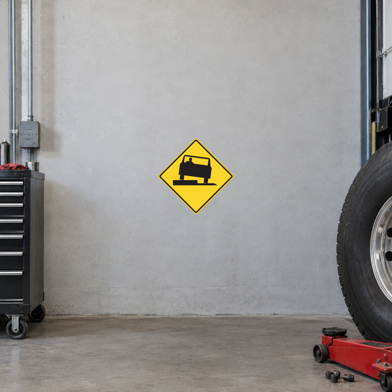 Road Sign Sticker applied in A truck inspection workshop, uninterrupted light grey painted concrete wall, heavy wheel at far right and low red jack below