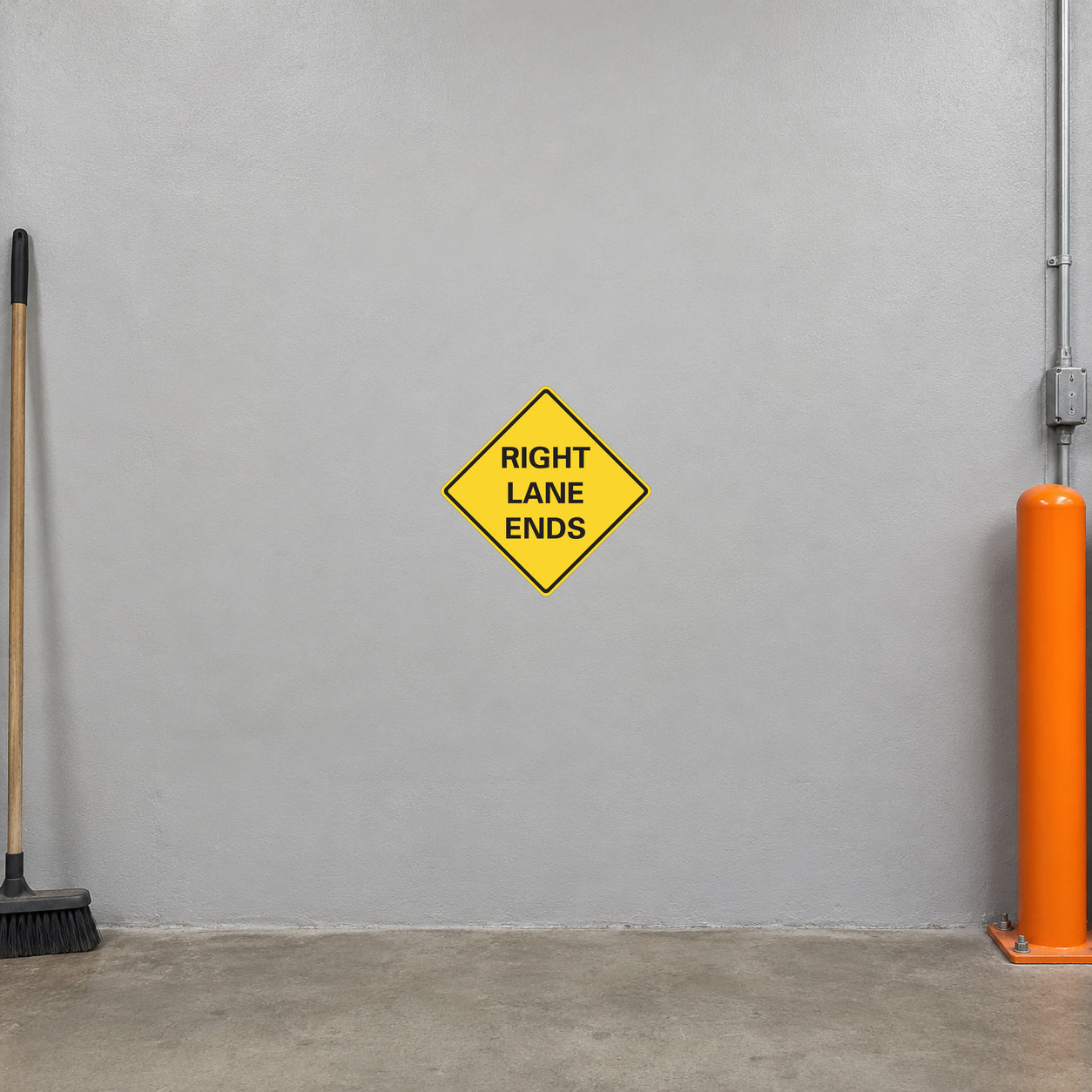 Road Sign Sticker applied in A municipal traffic supply workshop, one continuous smooth light grey painted concrete wall with absolutely no horizontal joints or door panels, orange bollard extreme right and broom extreme left, concrete floor below