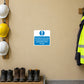 Safety Sign Sticker applied in a smooth pale ochre wall at a workshop PPE issue station, hard hats on hooks entirely at far left and safety boots at bottom below y90, centre clear