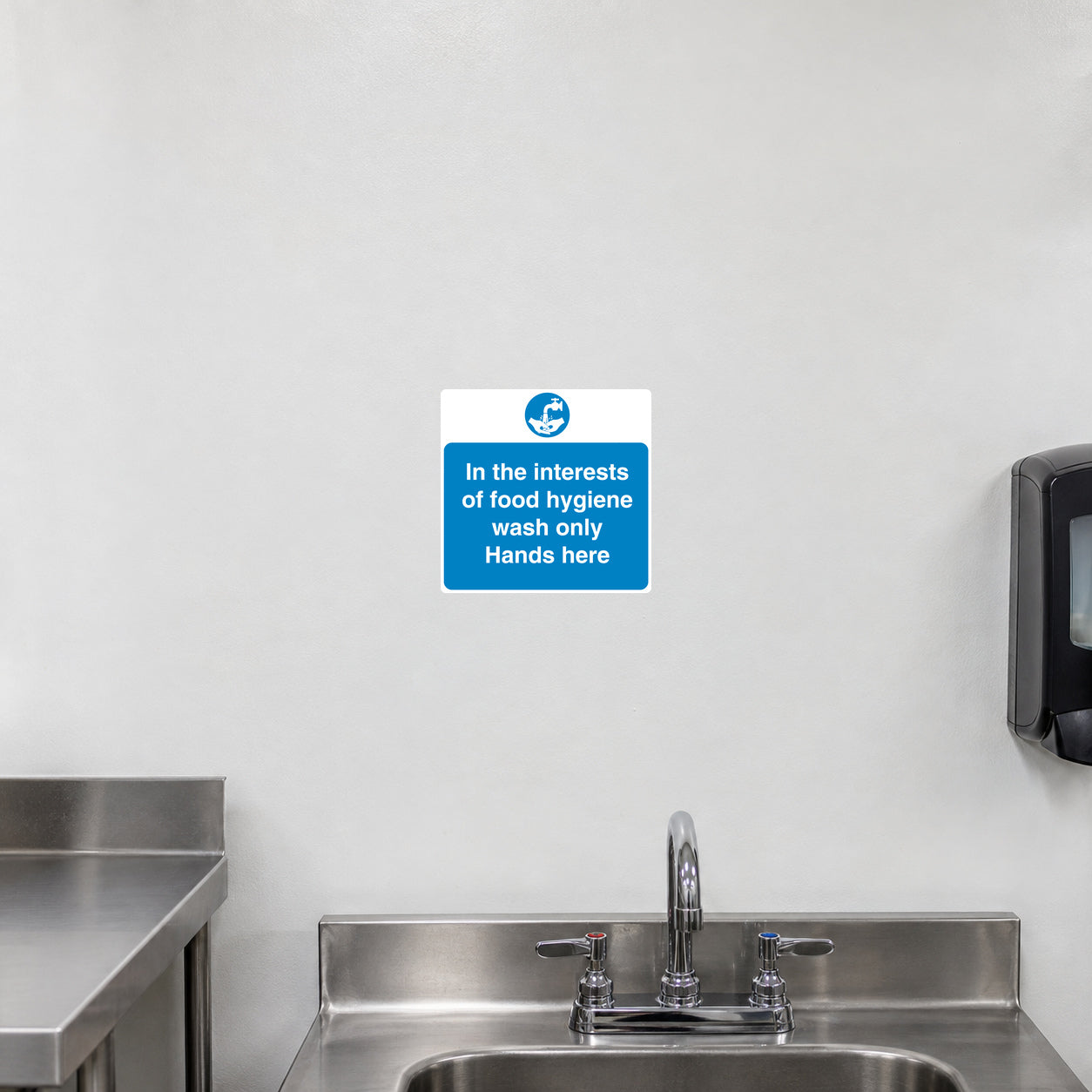 Safety Sign Sticker applied in a smooth white washable wall above a dedicated handwashing sink in a commercial food kitchen, sink and tap entirely below y85, soap dispenser far right and stainless bench extreme left