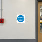 Safety Sign Sticker applied in a smooth cream wall next to a red manual alarm point in a school workshop corridor, alarm hardware far left and door frame far right, central blank surface