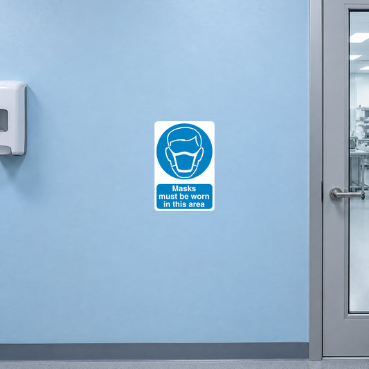 Safety Sign Sticker applied in a smooth light blue wall outside a clean assembly room, mask dispenser far left and glazed door frame extreme right