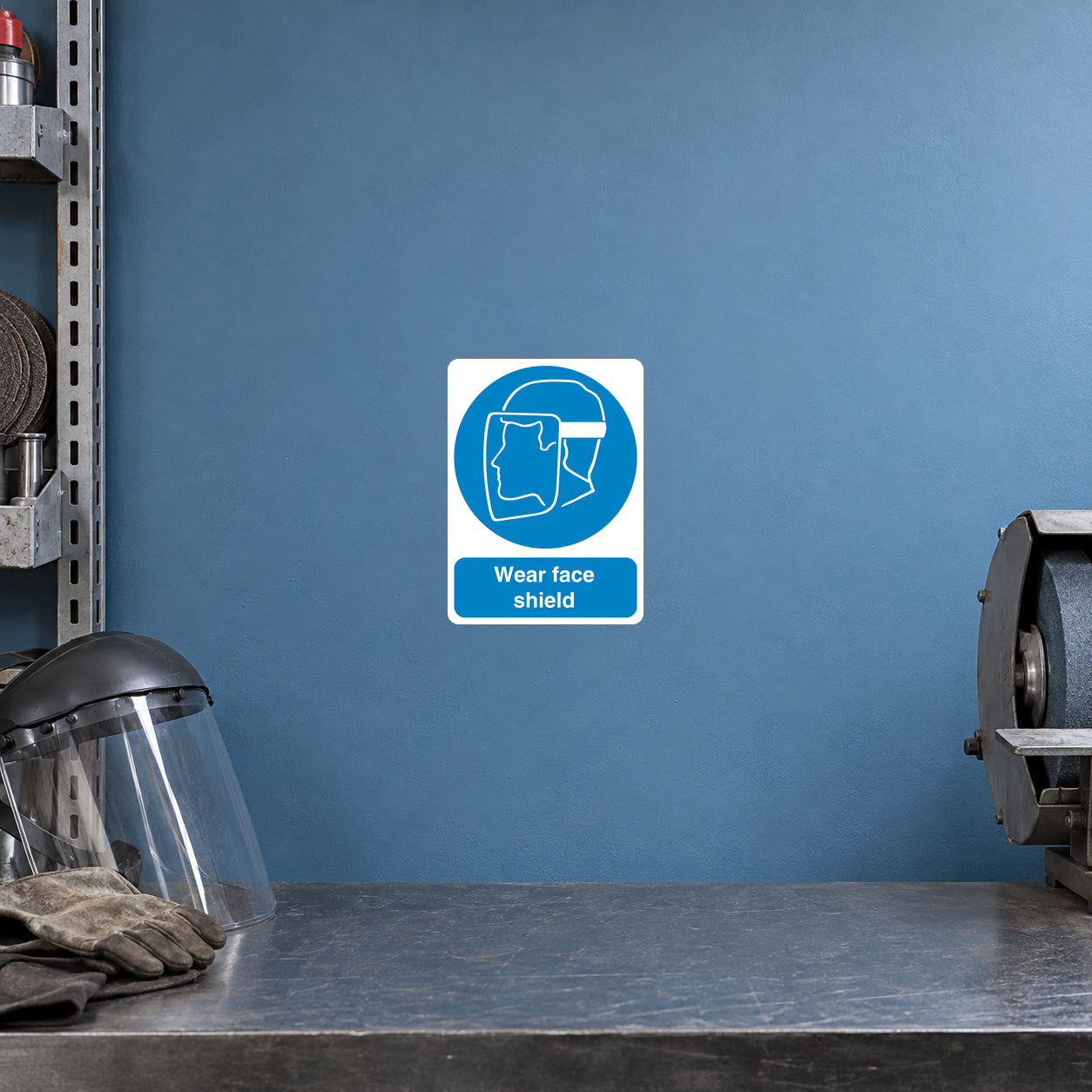 Safety Sign Sticker applied in a smooth muted blue wall at a metal grinding PPE station, face shield resting bottom left and grinder cropped far right