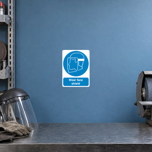 Safety Sign Sticker applied in a smooth muted blue wall at a metal grinding PPE station, face shield resting bottom left and grinder cropped far right