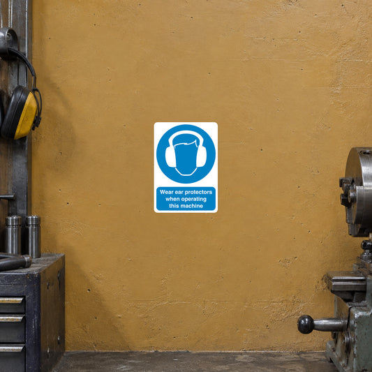 Safety Sign Sticker applied in Muted ochre factory wall beside a metal lathe, industrial hearing protectors hanging only at far left, lathe edge at far right, soft workshop daylight