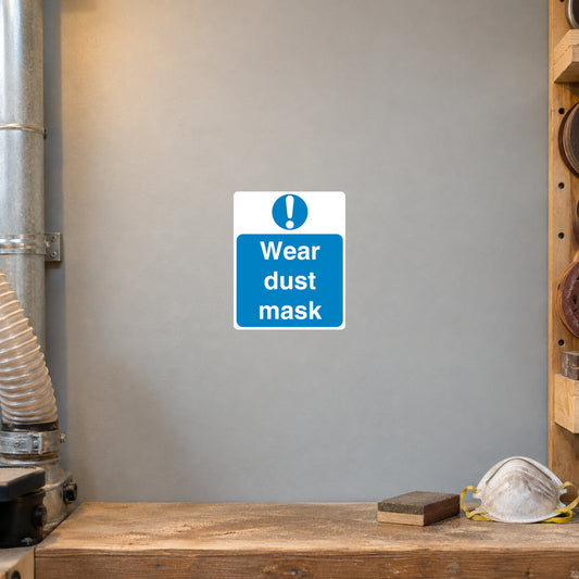 Safety Sign Sticker applied in Light grey smooth wall at a woodworking dust extraction station, dust mask and sanding block on a low shelf beneath y85%, warm sawmill light