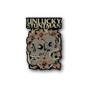 Vinyl sticker with Unlucky Stuntman text, twin skulls and leaves, tan.