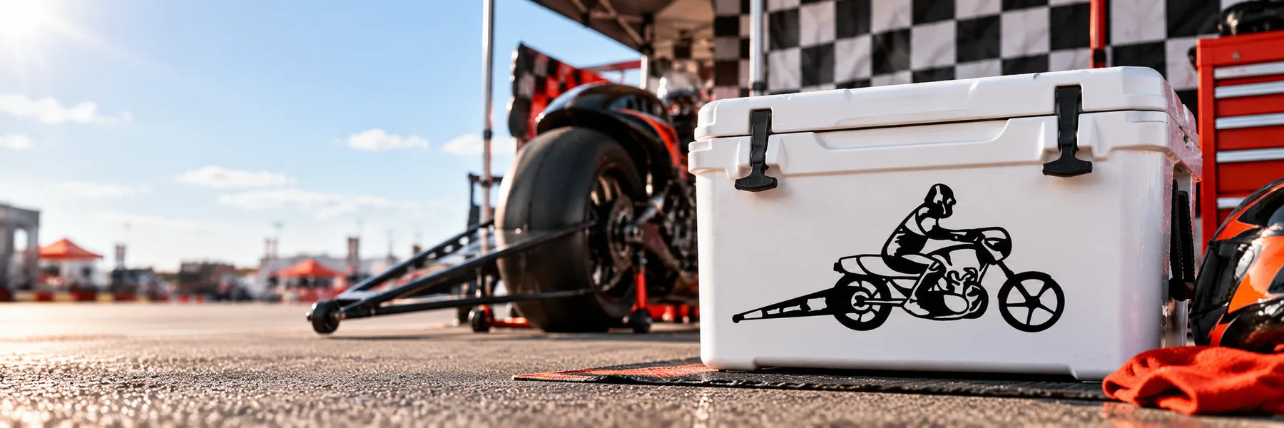 Illustrative Racing Decals application scene: White cooler in a sunlit motorcycle racing paddock, exact reference drag-racing silhouette in black vinyl on cooler front, race bike and checkered pit fabric behind, vivid orange/red accents, dynamic ground-level angle.